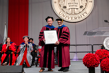 Stephen Lynch standing with Gregory both wearing a cap and gown standing on a stage