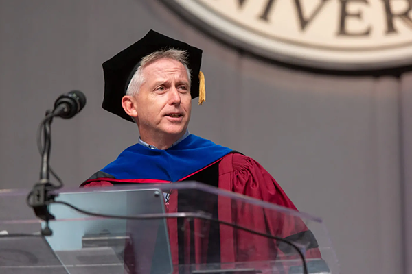 Stephen Lynch wearing a graduation hat and cap speaking at a pedestal