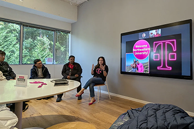 Three people sitting by a table. A tv is mounted on the wall to the right of the people, and it shows the T-Mobile logo. 